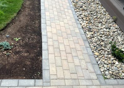 Curved walkway with patterned pavers, flanked by grass, pebbles, and plants.